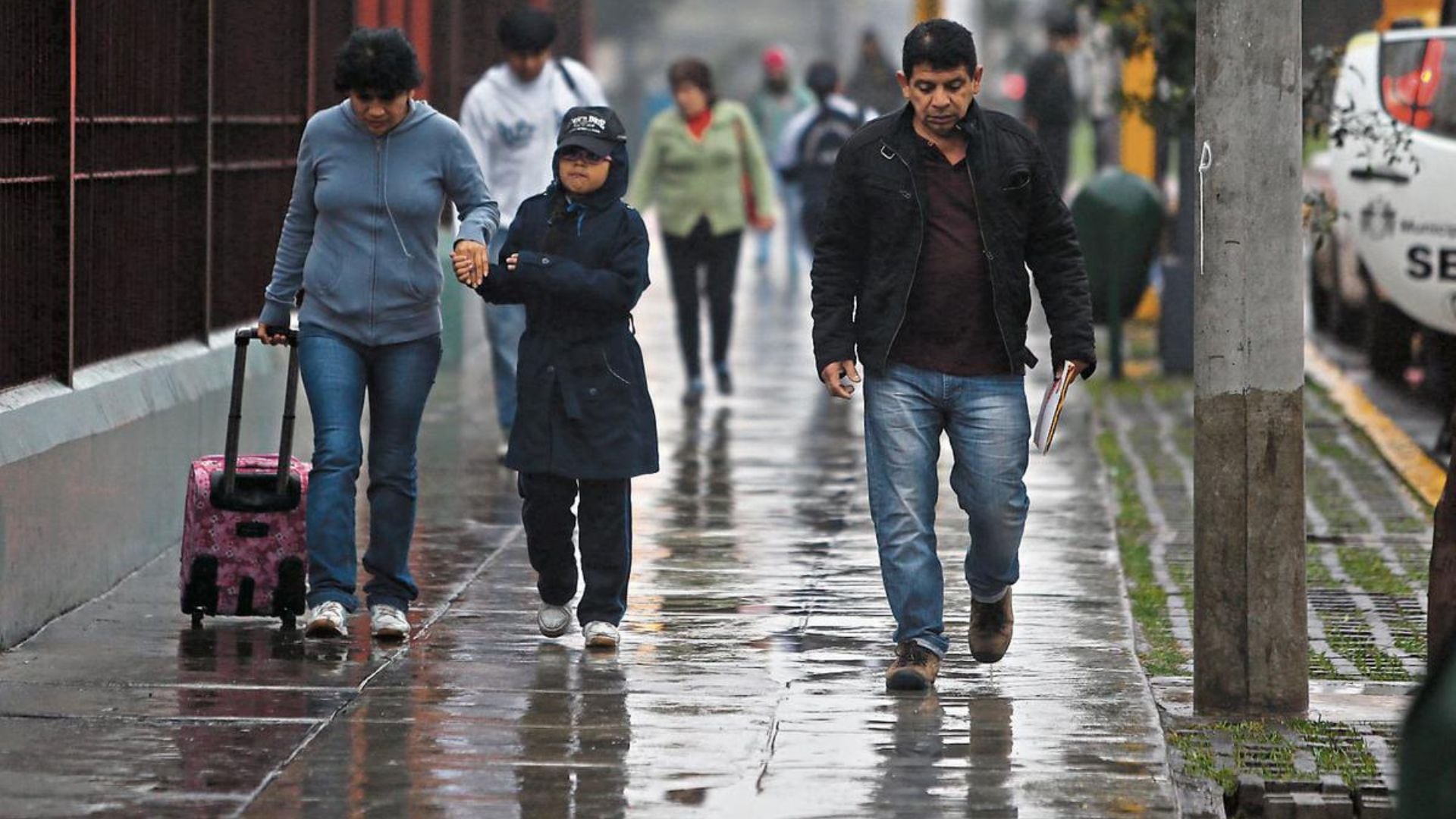 Una madre de familia y su pequeña hija, además de un hombre, caminan por la acera de una calle de Lima en pleno otoño y bajo una inesperada lluvia propia de la estación.