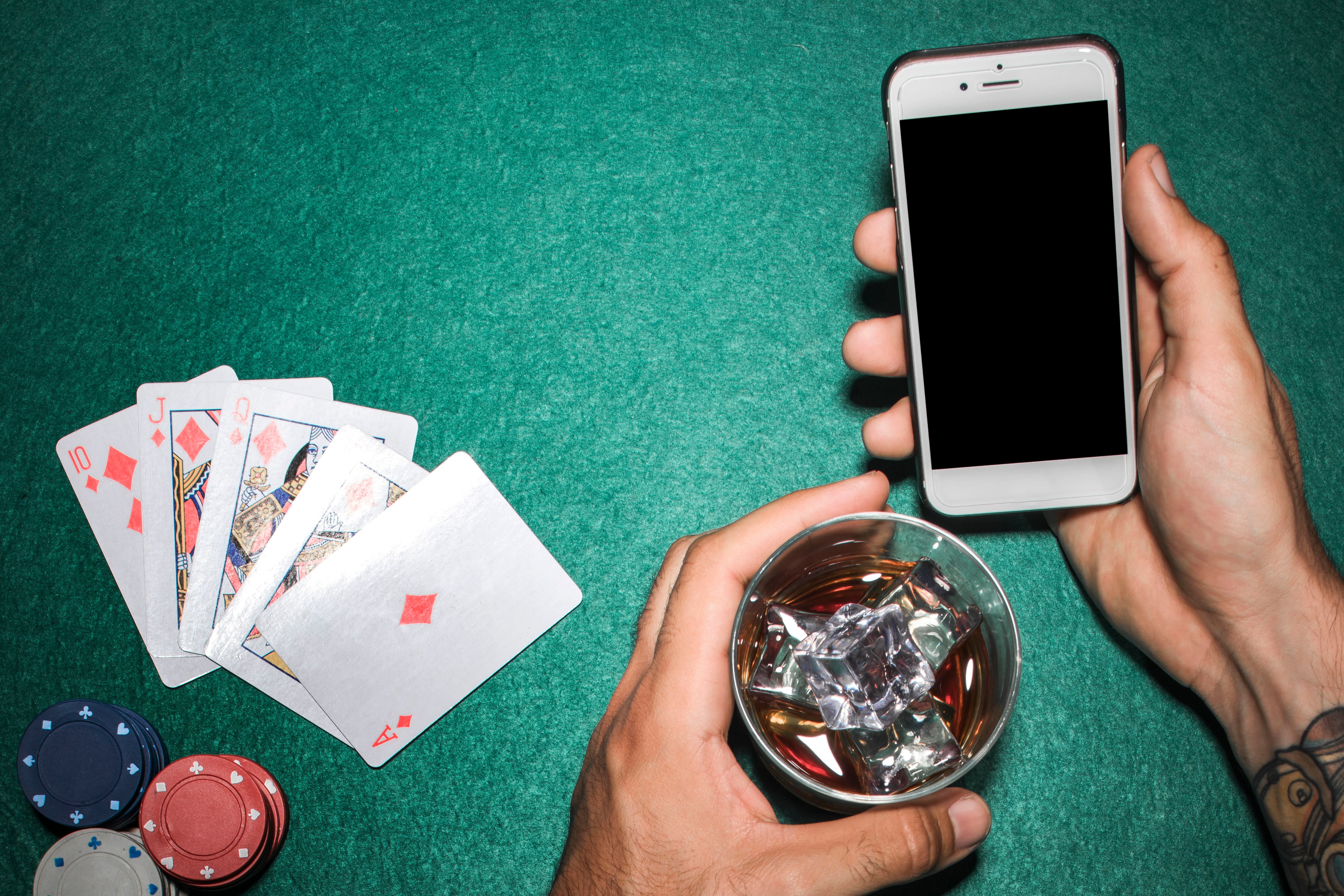 Hombre sujetando un móvil y una copa de alcohol junto a unas cartas y fichas de póker (Freepik)