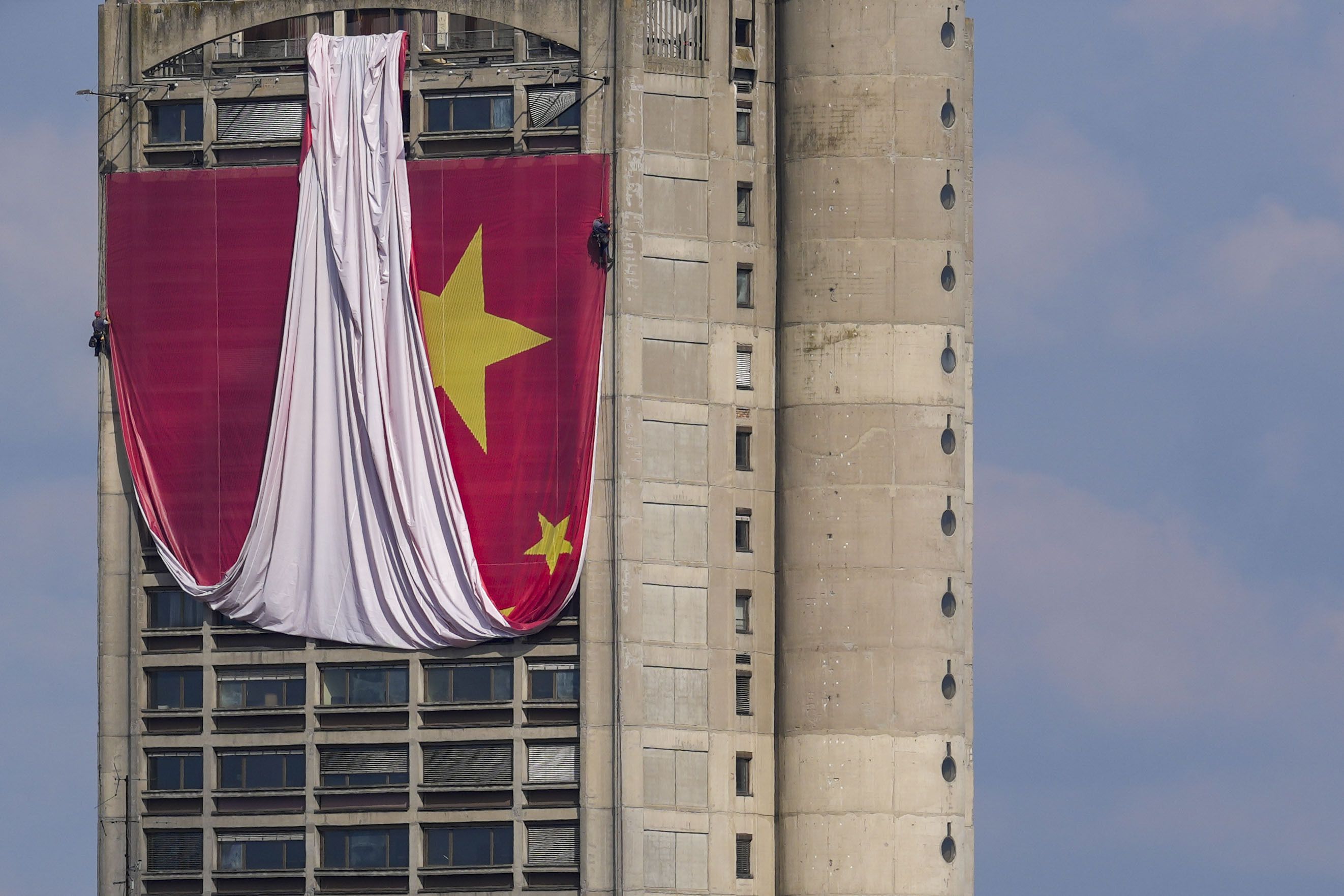 Trabajadores se descuelgan con cuerdas para instalar una bandera china gigante en un rascacielos que es un acceso simbólico a la ciudad desde el aeropuerto, en Belgrado, Serbia, el sábado 4 de mayo de 2024. (AP Foto/Darko Vojinovic)