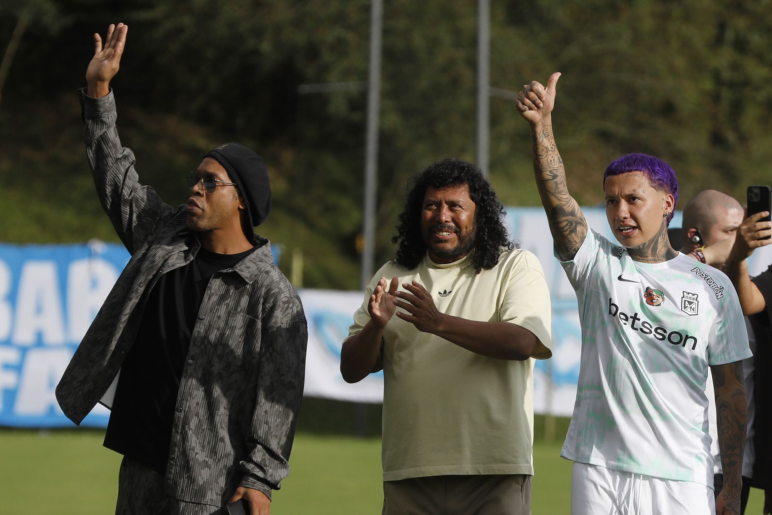 El cantante colombiano Blessd (d), junto a el astro brasileño Ronaldinho (i), y el exarquero colombiano, René Higuita, saludan a algunos simpatizantes durante una práctica de fútbol en la sede deportiva de Atlético Nacional - crédito Luis Eduardo Noriega Arboleda/EFE